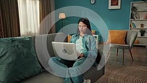 An attractive Asian woman sits on a sofa in the living room with a laptop on her lap. A woman is working, typing a text