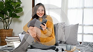 Attractive Asian woman in sweater and socks sitting in living room and using her smartphone