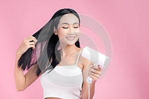 Attractive Asian woman usinf hair dryer on pink background