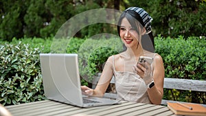 An attractive Asian woman working remotely in the garden, using her laptop computer and smartphone