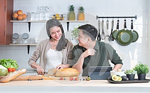 Attractive Asian young sweet couple cooking at home kitchen together. Beautiful wife smiling and slicing bread, enjoy preparing