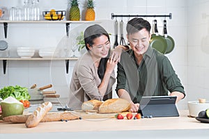 Attractive Asian young sweet couple looking recipe by using tablet at home kitchen, smiling, enjoy cooking preparing healthy