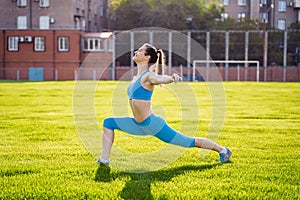 Attractive, athletic girl in a blue suit stretches in the stadium from green grass. Enthusiastically does a warm-up before the