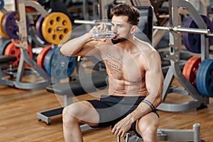 Attractive athletic young man bodybuilder fitness model drink water after exercise with barbell on the bench in gym.