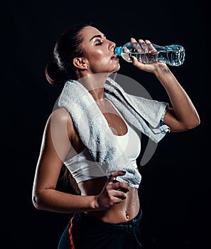 Attractive athletic young woman with perfect body drinking water from a bottle with towel around her neck against black background