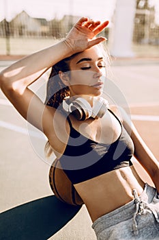 Attractive athletic young woman posing outdoors sitting on basketball court in the morning leaning on basketball. Fashion