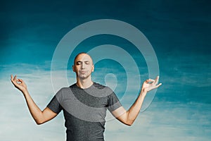 Attractive bald man with closed eyes and a calm face on a blue background holds his hands in a meditation pose.