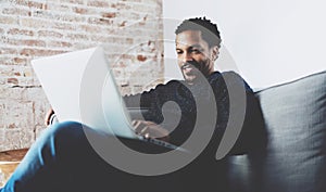 Attractive bearded African man smling and working on laptop while sitting sofa at his modern home office.Concept of