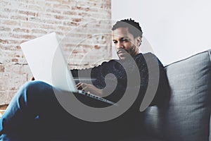 Attractive bearded African man working on laptop while sitting sofa at his modern home office.Concept of young people