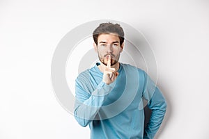 Attractive bearded man asking to keep quiet, showing taboo hush gesture and looking at camera calm, standing over white