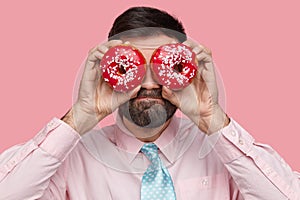 Attractive bearded man carries doughnuts near eyes, has dark stubble, dressed in formal outfit, hides face with