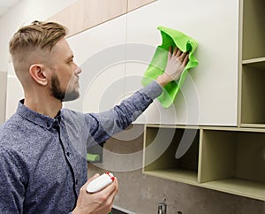 Attractive bearded man cleaning the handleless cabinet doors with a green rag in a modern kitchen