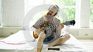 Attractive bearded man in headphones sitting on bed learning to play guitar using tablet computer in modern bedroom