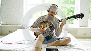 Attractive bearded man sitting on bed learning to play guitar using tablet computer in modern bedroom at home