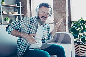 Attractive, bearded man sitting on the couch in living room, having headphones on his head, listening his favorite music, singing