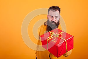 Attractive bearded man wearing yellow hoodie shoing red gift box over yellow background.