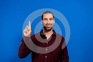 Attractive bearded spanish man in purple shirt pointing smiling up with his finger isolated on blue studio background