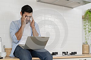 Attractive bearded young man use laptop computer to surfing social media or checking work while sitting on kitchen counter at home