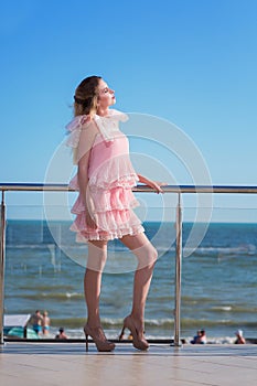 An attractive and beautiful female in a pink dress is posing on a sunny hotel`s balcony. Luxurious lifestyle.