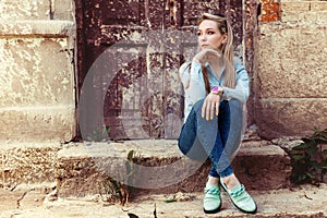 Attractive beautiful gentle girl sits in the city on the steps of the old building in jeans and fashion footwear