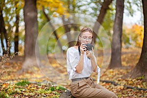 Attractive beautiful young girl holding modern mirror-less camera in autumn park