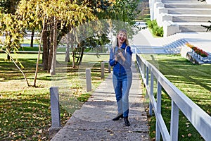 Attractive beautiful young girl holding modern mirror camera in autumn park. Traveller walking in place with yellow leaves on tree