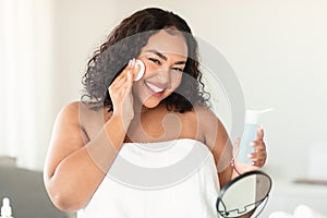 Attractive black body positive woman using facial toner and cotton pad for moisturising skin, making beauty treatments