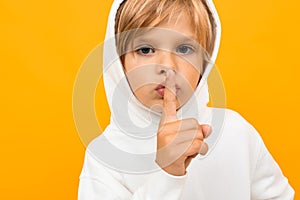 Attractive blond boy in a white hoodie asks to be quieter on an orange background