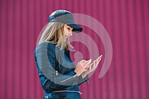 Attractive blond girl in leather black jacket and cap, surfing the Internet on a mobile phone