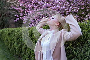 Attractive blond woman in stylish beige coat walks in spring park and straightens her hair. Happy woman outdoors