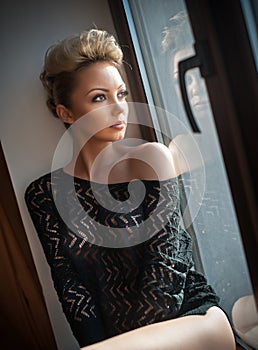 Attractive blonde with black see through blouse looking on the window in daylight. Portrait of sensual short fair hair woman