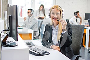 Attractive blonde businesswoman with headset sitting near computer and looking at camera in office.