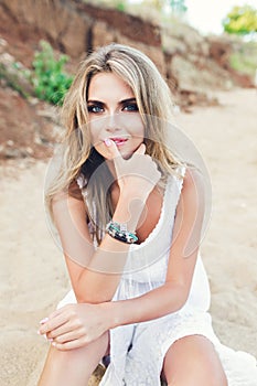 Attractive blonde girl with long hair and blue eyes is sitting on beach. She wears white dress, ornamentation. She is