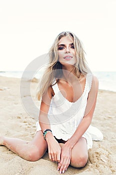 Attractive blonde girl with long hair is sitting on sand on beach. She wears white dress, ornamentation. She is looking