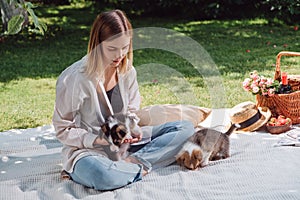 Attractive blonde girl sitting on white blanket in garden and having picnic at sunny day with cute puppies.