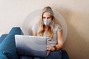 Attractive blonde girl wearing a protective mask works hard and responsibly with a laptop remotely at home. Isolated due to the