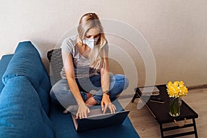 Attractive blonde girl wearing a protective mask works hard and responsibly with a laptop remotely at home. Isolated due to the