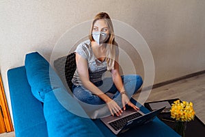 Attractive blonde girl wearing a protective mask works hard and responsibly with a laptop remotely at home. Isolated due to the