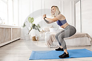 Attractive blonde-hair sportive woman is standing on blue mat, doing squats exercise. Young beautiful female is working