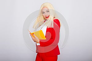 Attractive blonde haired woman wearing red jacket and pants holding orange book and pen
