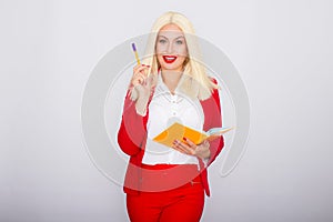 Attractive blonde haired woman wearing red jacket and pants holding orange book and pen