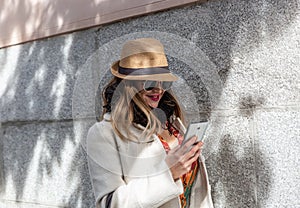 Attractive blonde with hat and black glasses looks smiling and happy at her smartphone