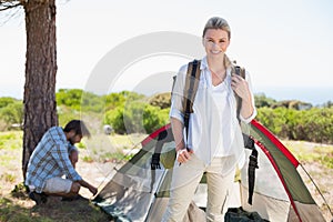 Attractive blonde smiling at camera while partner pitches tent