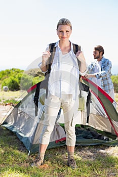 Attractive blonde smiling at camera while partner pitches tent