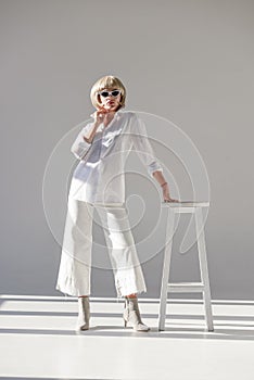 attractive blonde woman in sunglasses and fashionable white outfit leaning on chair