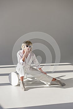 attractive blonde woman in sunglasses and fashionable white outfit sitting on chair