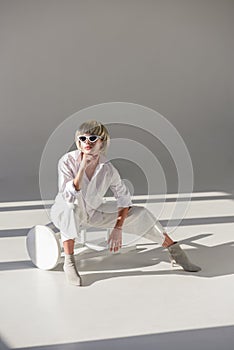 attractive blonde woman in sunglasses and fashionable white outfit sitting on chair and resting chin on hand photo