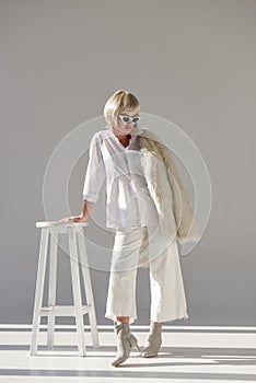 attractive blonde woman in sunglasses and fashionable winter outfit with faux fur coat leaning on chair