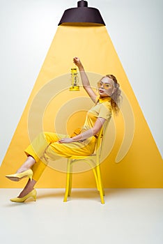 Blonde woman in sunglasses sitting on chair and holding vintage lamp on white and yellow