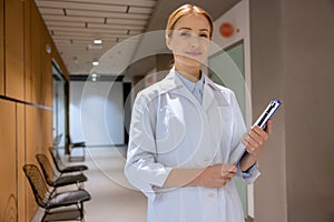 Attractive blonde young female doctor standing in hospital corridor and holding clipboard.
